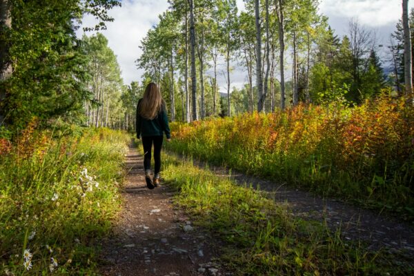 woman in black jacket walking on pathway between green grass and trees during daytime