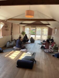A small group of people in a yoga studio sitting facing one another, on yoga mats and cushions with sunlight coming in from a window to the side and a door at the end of the room looking out into a garden.
